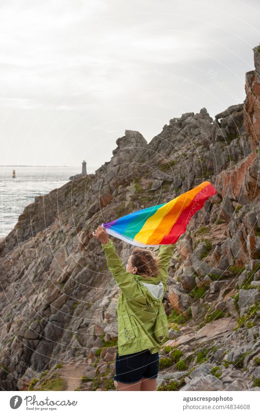 Blonde woman raising her arms at the tip of cape waving a rainbow flag, in the background a rock wall and a lighthouse in the sea. LGBTQ unrecognizable lesbian