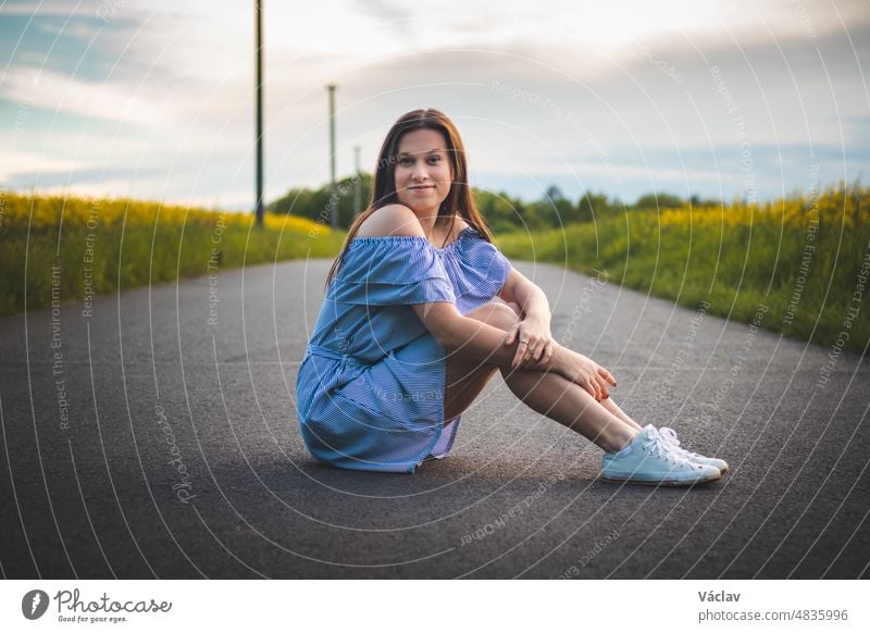 Candid portrait of a shy young girl in a beautiful blue summer dress with white sneakers sitting on the road with a natural breathtaking smile at sunset. Fashion style. European model
