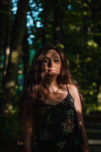 pensive view of a 24 year old woman with dark brown hair in a forest at sunset. Candid portrait of handsome woman in summer dress. Hidden in the shadows