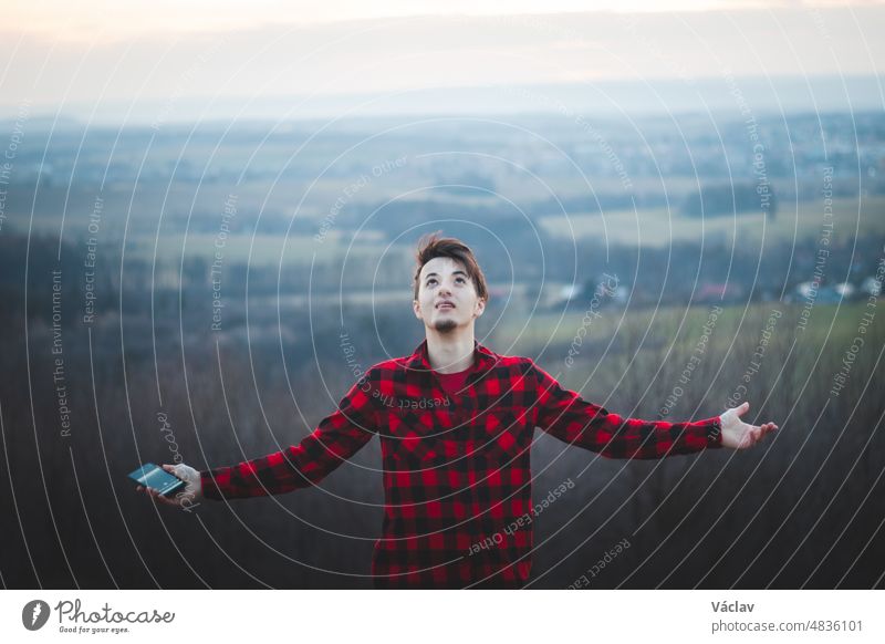Candid portrait of a man in his 20s dressed in a black and red checked shirt joyfully facing the sky. He gives thanks to be on earth. Authentic people at their emotions