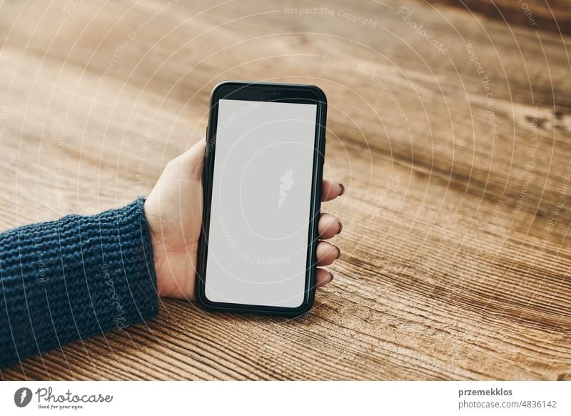 Female hand holding smartphone mockup with solid white color blank empty isolated screen on wooden background. Person using technology. Mobile phone with copy space left