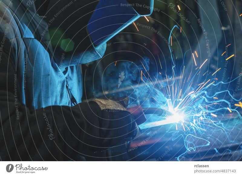 strong man is a welder, in a welding mask and welders leathers, a metal product is welded with a welding machine in the garage, blue sparks fly to the sides