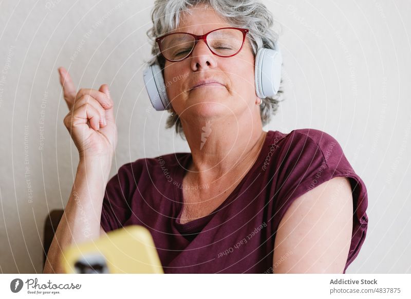 Happy mature woman listening to song in headphones via smartphone music sing positive meloman using playlist happy eyes closed female senior casual eyeglasses