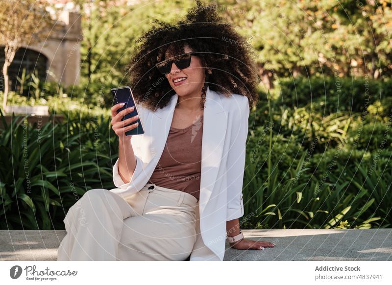 Hispanic businesswoman browsing on cellphone on street smartphone urban female message hispanic ethnic sunglasses smart casual text bench fence sunny style
