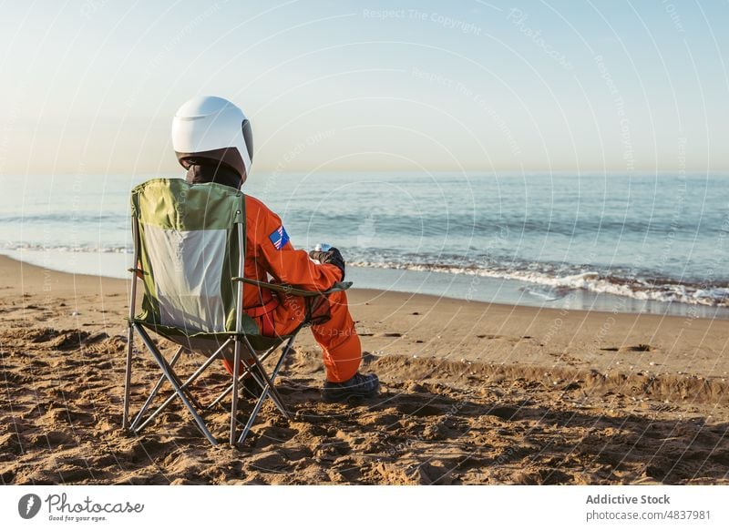 Spaceman sitting on chair and admiring sea spaceman beach astronaut admire evening rest concept futuristic resort sand vacation spacesuit helmet cosmonaut relax