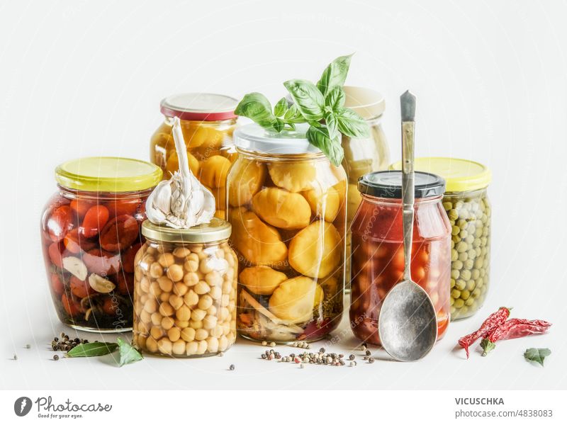 Preserved vegetables selection in glass jars with herbs, spices and spoon at white background: conserved chickpeas, zucchini, beans and tomatoes. preserved