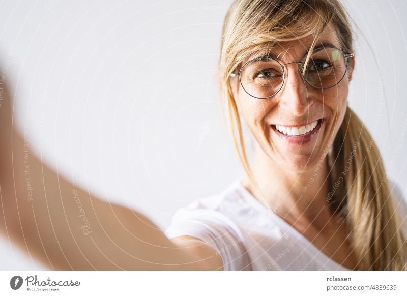 Attractive blonde woman dressed casually and glasses look smiling broadly. Beautiful woman having cheerful expression while posing against white background, stretching arm to camera for a selfie