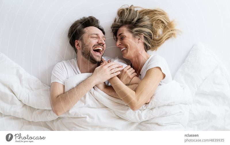 Top view of young loving couple lying on bed laughing and smiling with blanket. They are looking at each other with trust in love man happy home intimate