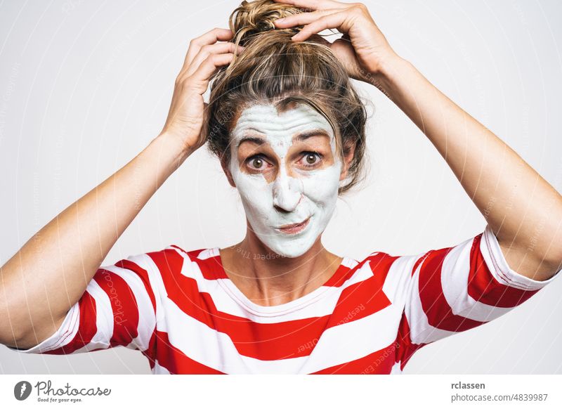Cheerful woman correcting hair bun, has clay mask on face, enjoys softness, has beauty treatments at home, on gray background. Skin care concept image skin