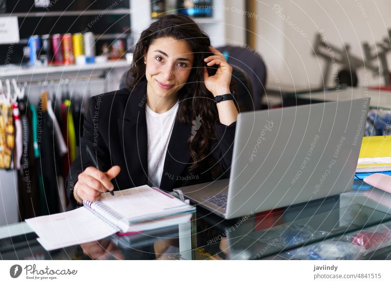 Happy sports psychologist looking at camera woman write notebook gym smile psychology session portrait female work touch hair brunette desk young laptop