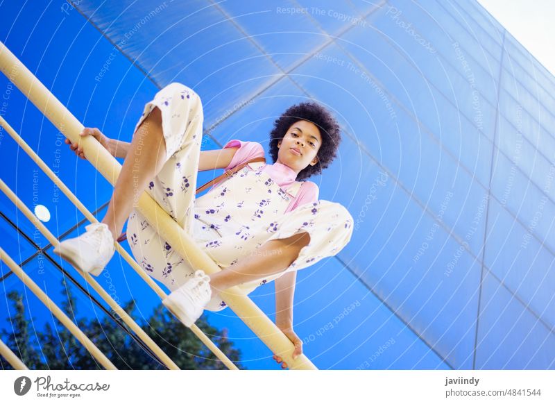 African American woman in casual clothes, sitting on a railing of a modern building. positive looking at camera happiness afro hairstyle black curly person girl