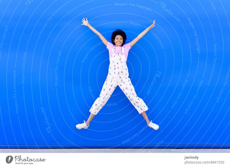 African American female jumping with raised arms against a blue urban wall. woman happiness joy happy smiling smile afro hairstyle black curly person girl
