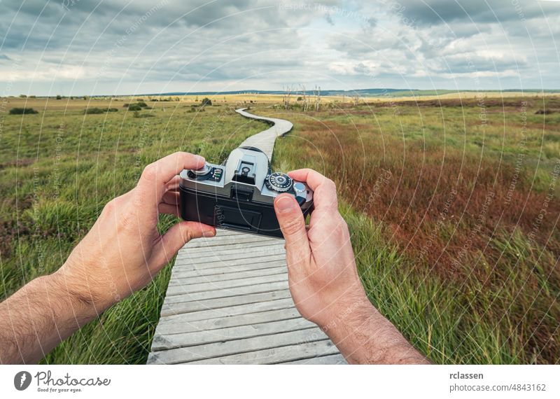male hand holding a vintage camera against the a landscape with boardwalk to take a picture, pov, point of view perspective. man people lifestyle adventure
