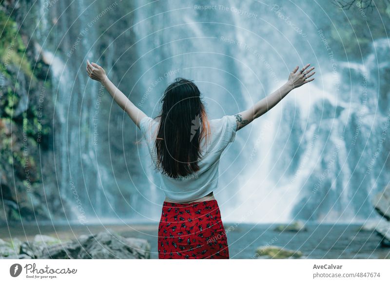Young woman jumping, celebrating and expressing happiness in front of a waterfall, traveling and touring alone. freedom in nature, independent lifestyle and freedom concept. Back image