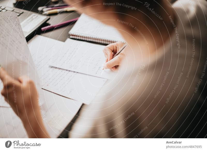 Close up detail image of a young woman studying and working on his home desk, doing homework during university, preparing for exam with textbook and taking notes, selective focus on the pencil.