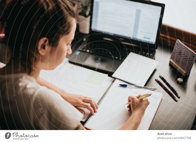 Top view of the back of a young woman studying and working on his home desk, doing homework during university, preparing for exam with textbook and taking notes, selective focus on the pencil.