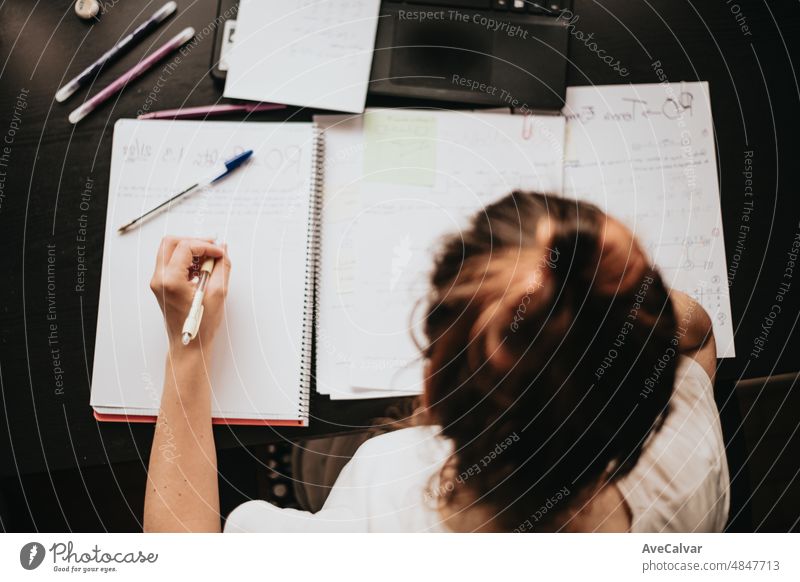 Top view of the back of a young woman studying and working on his home desk, doing homework during university, preparing for exam with textbook and taking notes, selective focus on the pencil.