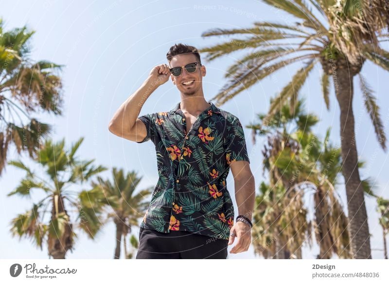College student wearing flowered shirt, black shorts, sunglasses, walking on walkway young man smiling palms low angle toothly smile looking at camera person
