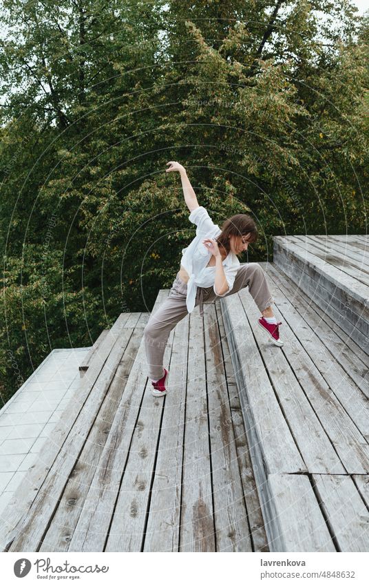 Young female in white shirt and grey linen pants dancing on the stairs in front of the foliage, woman dancer choreographer outdoors girl stylish urban movement