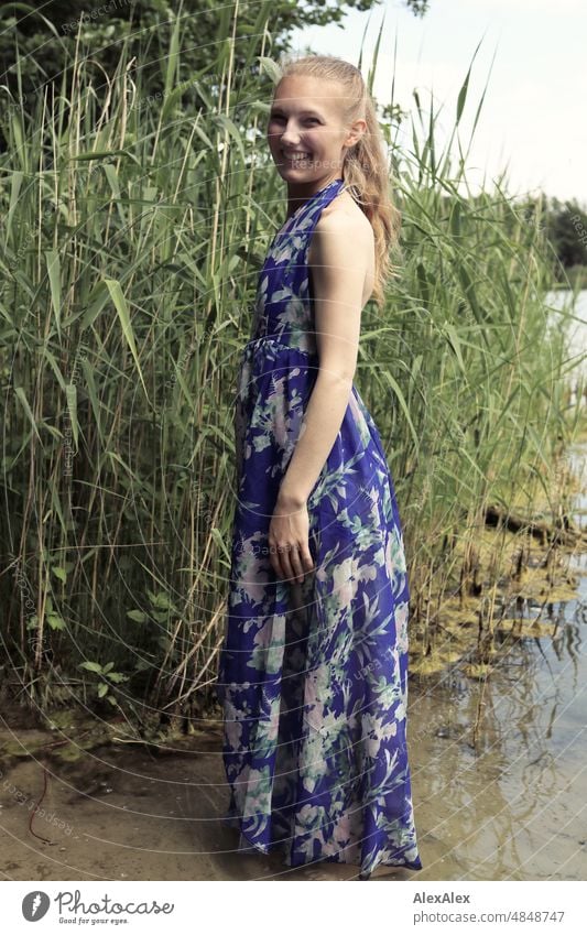 Young red blonde long haired woman stands in dress with feet in lake on shore and smiles sideways at camera Woman youthful pretty Slim Grass Nature Blonde