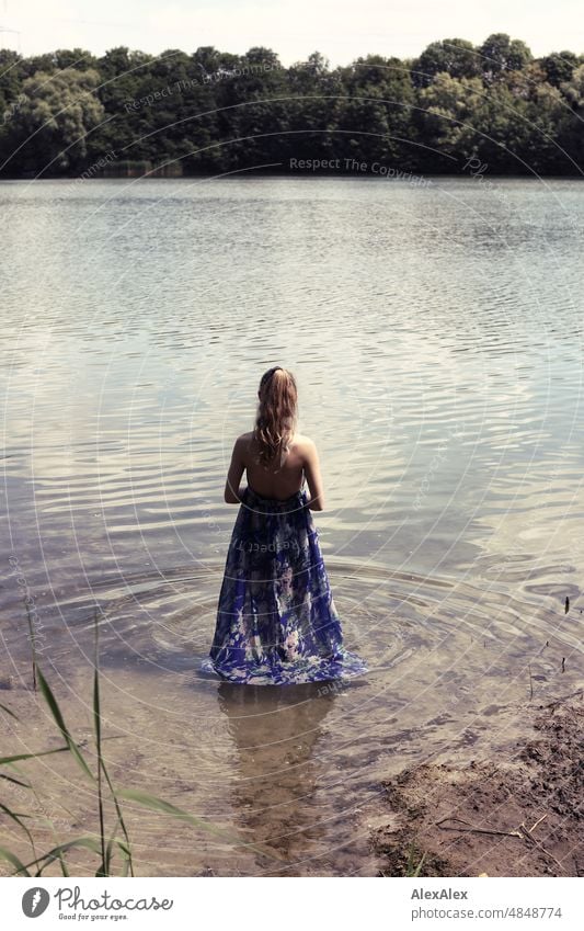 Young red blonde long haired woman in dress standing with feet in lake on shore with back to camera Woman youthful pretty Slim Grass Nature Blonde cheerful