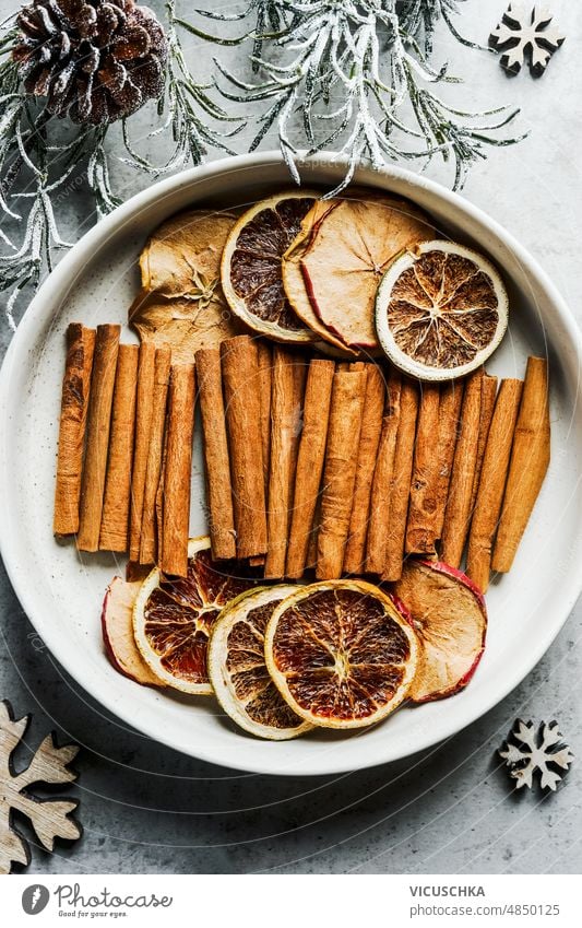 Dried citrus slices and cinnamon sticks in white bowl with fir green and pine cones. Winter flavor dried winter fruits spices top view background dried orange