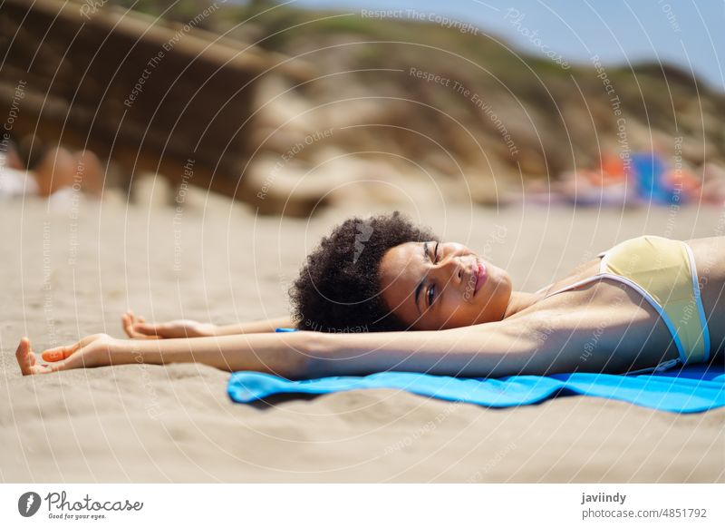 Content young African American woman lying on sandy beach and winking at camera sunbath relax arms raised vacation seashore holiday enjoy summer traveler female