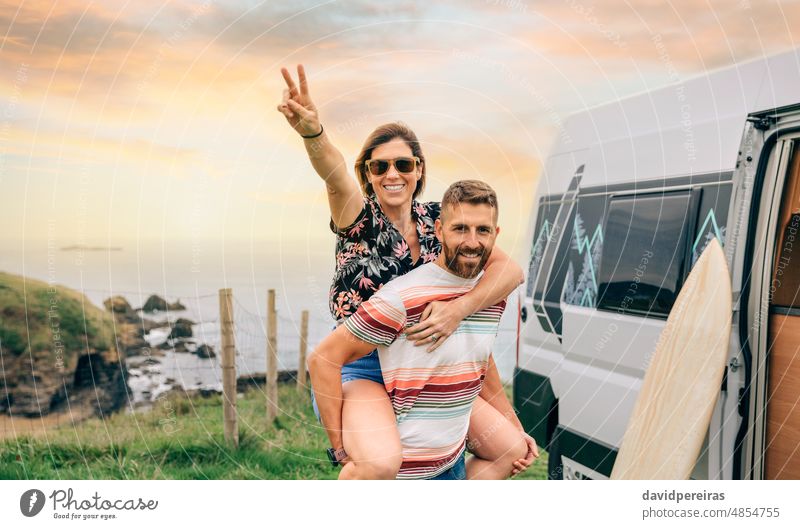 Couple looking camera having fun piggybacking next to their camper van young couple happy trip victory sign v sign hand smiling enjoy sunglasses arm woman