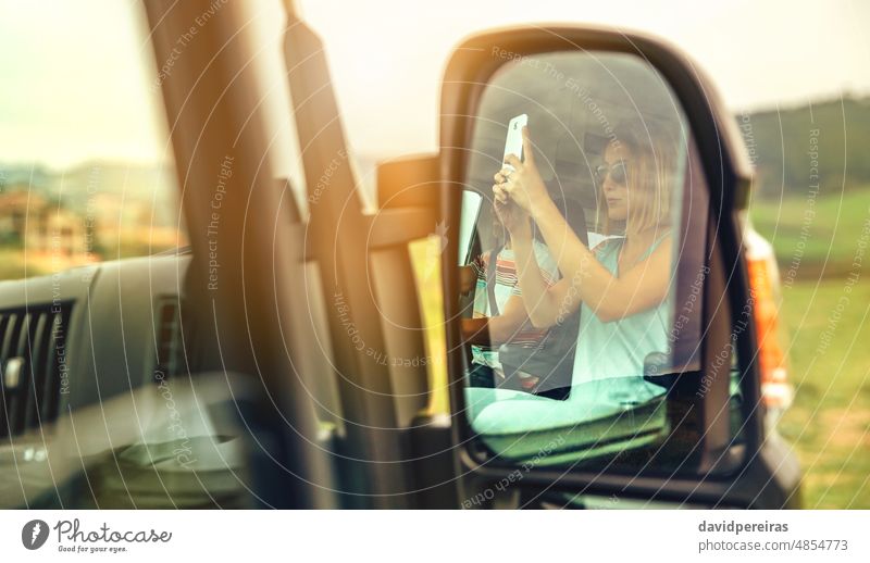 Reflection in a camper van rear-view mirror of a woman taking a picture with her cell phone reflection friend driving mobile sunglasses sitting summer trip