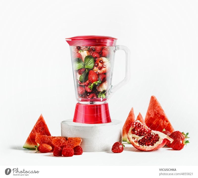 Glass blender filled with red fruits: watermelon slices, pomegranate seeds, raspberries, strawberries and grapefruit on product podium at white background with ingredients.