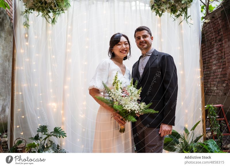 Newlywed multiethnic couple with bouquet of flowers newlywed bride groom hug bloom wedding arch multiracial diverse asian blossom green curtain garland tuxedo