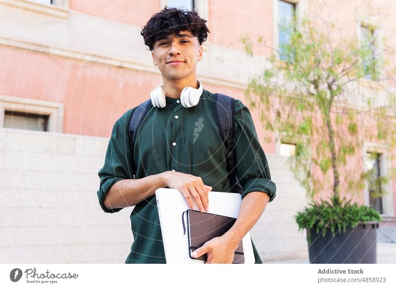 Smiling ethnic man with laptop on street student netbook headphones city building summer lifestyle smile happy delight cheerful joy carefree notepad planner
