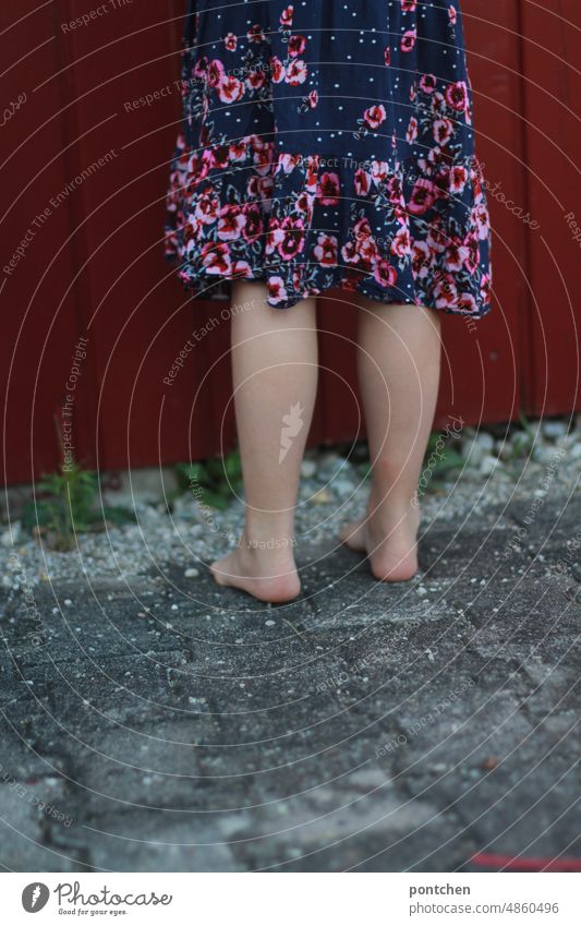 Children's game. Hide lower body. Averted child in dress stands in front of a red wooden hut, hut. Stand Dress averted turn away Playing Barefoot look away view
