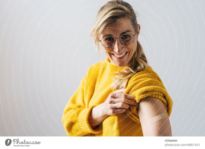 Portrait of a mature woman with glasses smiling after getting a corona vaccine. Woman holding up her shirt sleeve and showing her arm with bandage after receiving vaccination.