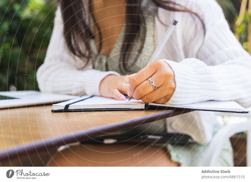 Anonymous self employed woman writing in notebook in outdoor cafe take note write freelance plan schedule terrace work laptop street modern summer asian ethnic