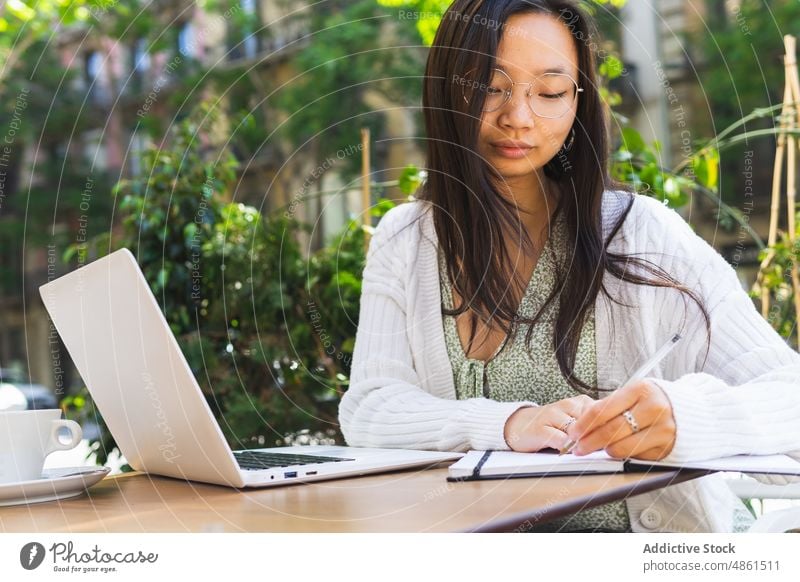 Asian self employed woman writing in notebook in outdoor cafe take note write freelance plan schedule terrace work laptop street city modern summer asian ethnic