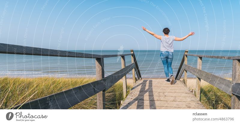 woman is raising hands on a wooden path to the Baltic sea over sand dunes beach walkway raised boardwalk travel adventure efficiency energy baltic north sea