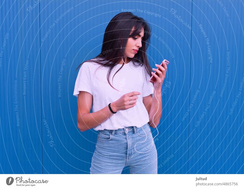 Young girl of Arab ethnicity using her cell phone in front of a blue wall Girl Ethnicity spain Woman Twenties Trendy BuildingExterior Young Female Young Adults
