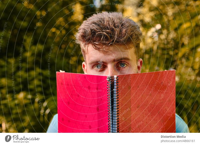 Blue-eyed student scared about exams covering his face with a red notebook outdoors autodidact autumn beautiful beauty caucasian culture cute education