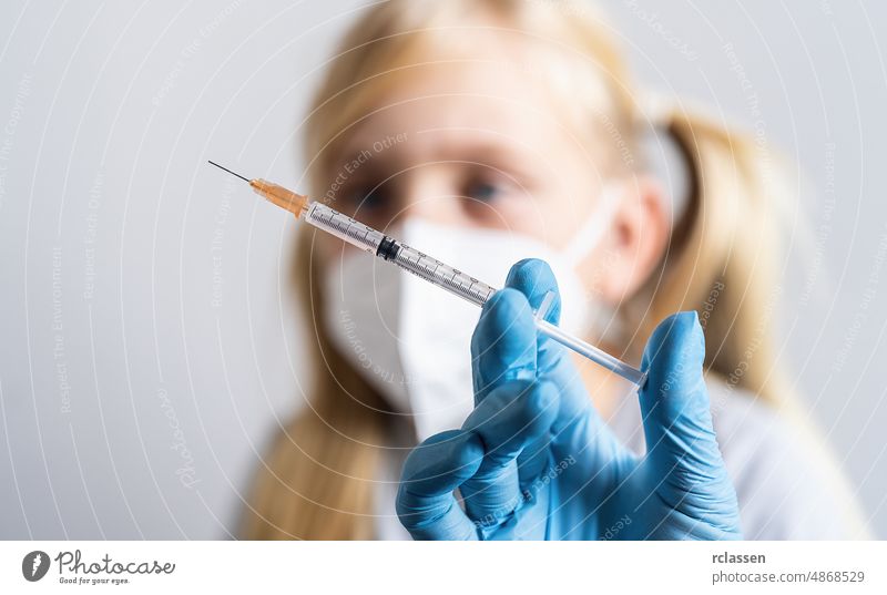 Close-up medical syringe in doctors hand with vaccine for covid-19 coronavirus, flu, and blond child in background school dangerous injection shot pandemic