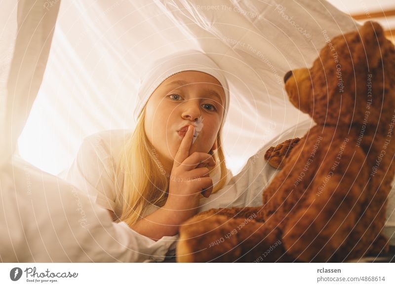 Sweet little girl with a teddy bear hold finger on lips showing silence to keep a secret while lies on her bed in a in tent with nightcap. child kid story toy