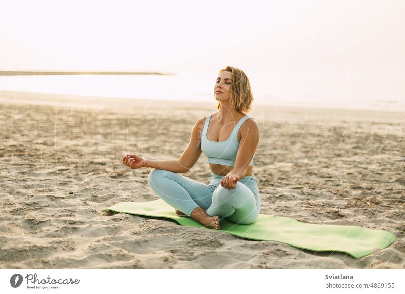 A happy young woman is sitting in a lotus position and doing yoga on the beach at sunset. The concept of a healthy active lifestyle sea beautiful female fitness