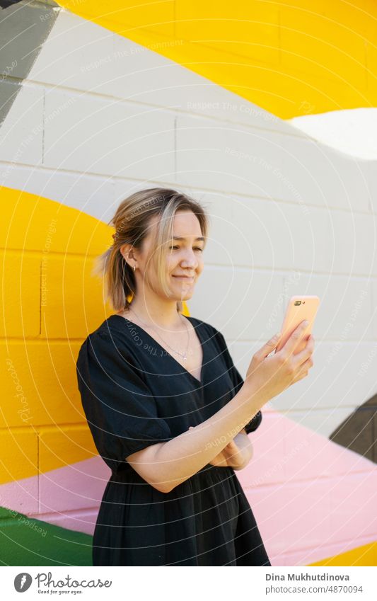 Young woman using mobile phone standing near colorful wall taking selfie or video chatting on smartphone. Screen time. Lifestyle using technology. Young remote worker employee in modern office space.