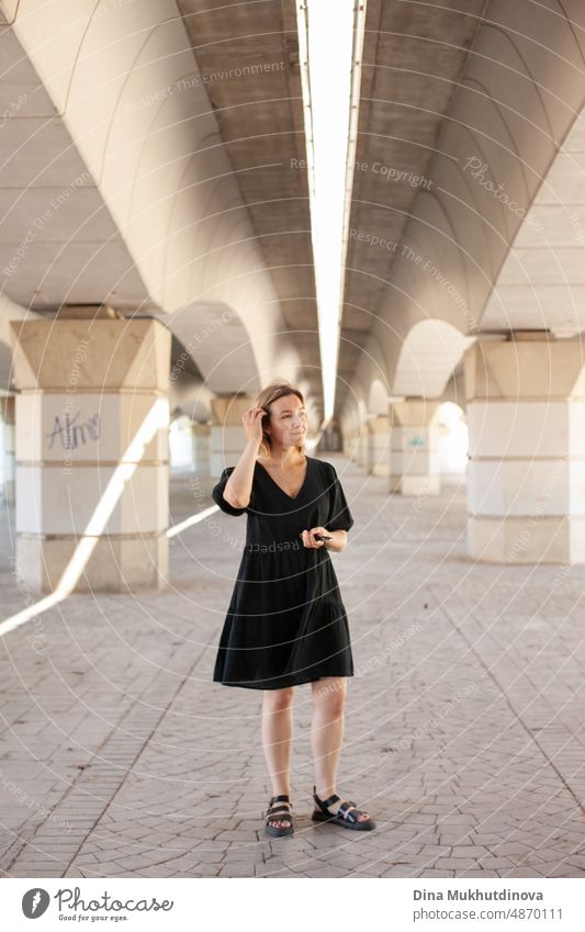 Young woman in casual black dress walking under the bridge in urban location in the city. Young generation Z style photoshoot. Real people lifestyle in the city. Geometry in city architecture. Woman walking towards the camera.