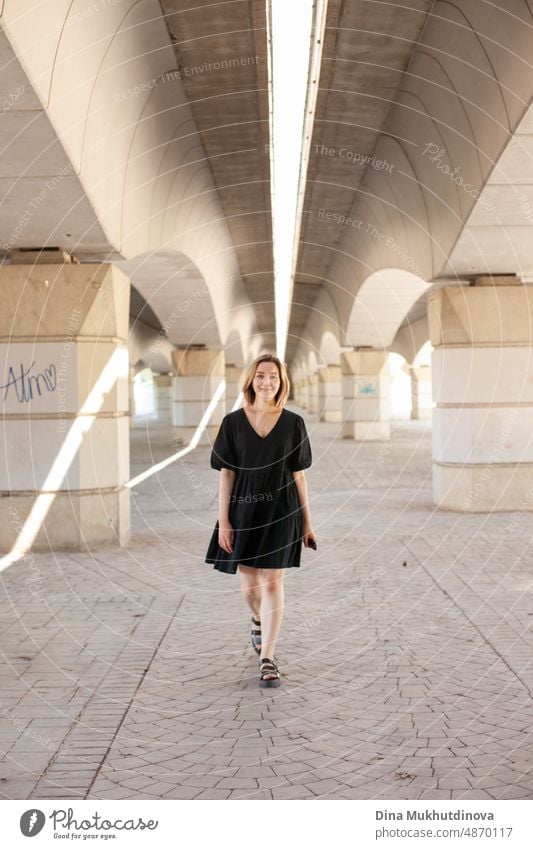 Young woman in casual black dress walking under the bridge in urban location in the city. Young generation Z style photoshoot. Real people lifestyle in the city. Geometry in city architecture. Woman walking towards the camera.