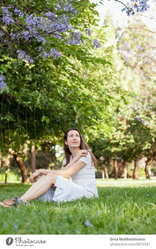 Young woman brunette sitting in the park on the green grass in summer. Lifestyle candid photo outdoors of woman dressed in casual clothes. Relaxing moments in the park in spring.