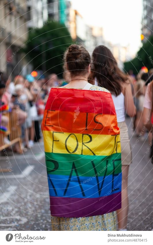 Woman from behind walking in rainbow Pride flag with words: Love is only for the brave. LGBTQIA+ support and ally. Pride parade in the streets of European city.