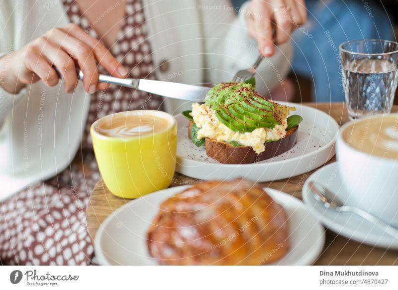 Eating avocado toast with knife on wooden table with pastries, glass of water and coffee cup. Drinking cappuccino at brunch with friends. Candid lifestyle with food and drink. Millennial way of living. View from above on the table with plates and meals.