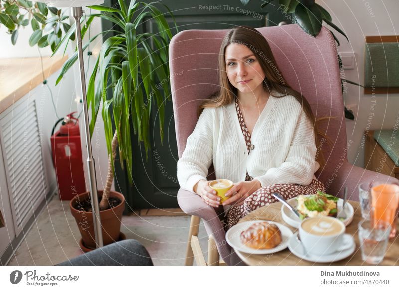Beautiful woman drinking coffee at cafe sitting in pink chair. Candid millennial lifestyle. Attractive face of an adult woman in her thirties. Looking out the window at restaurant, holding cup of coffee. Coffee break in cafeteria at work in modern office.
