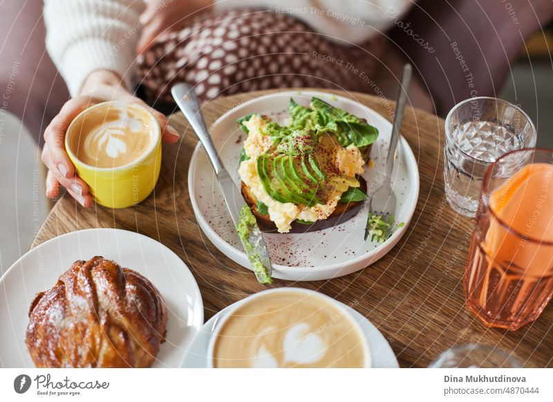 Avocado toast on wooden table with pastries, glass of water and coffee cup. Drinking cappuccino at brunch with friends. Candid lifestyle with food and drink. Millennial way of living. View from above on the table with plates and meals.